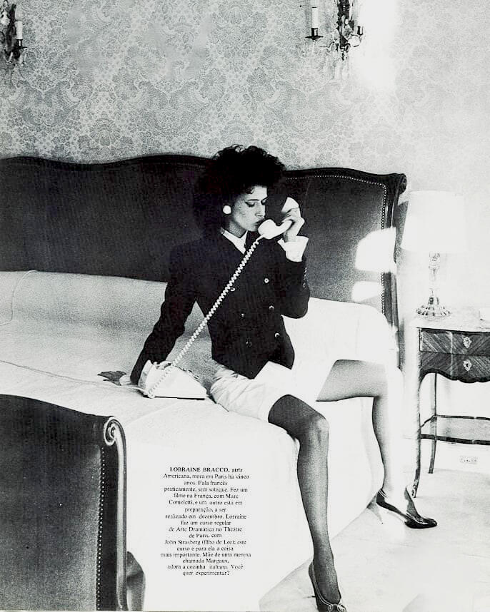 Black and white photograph from Interview Brazil magazine featuring actress Lorraine Bracco. She is sitting on the edge of a bed in a YSL dark blazer and heels, holding a vintage telephone receiver to her ear.