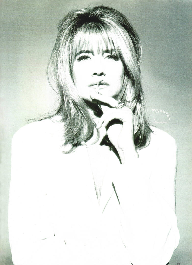 Vogue Germany magazine spread featuring a high-contrast, black and white fashion portrait of actress Lorraine Bracco with blonde hair and bangs. She is wearing a white blazer and posing thoughtfully with her hand near her chin.