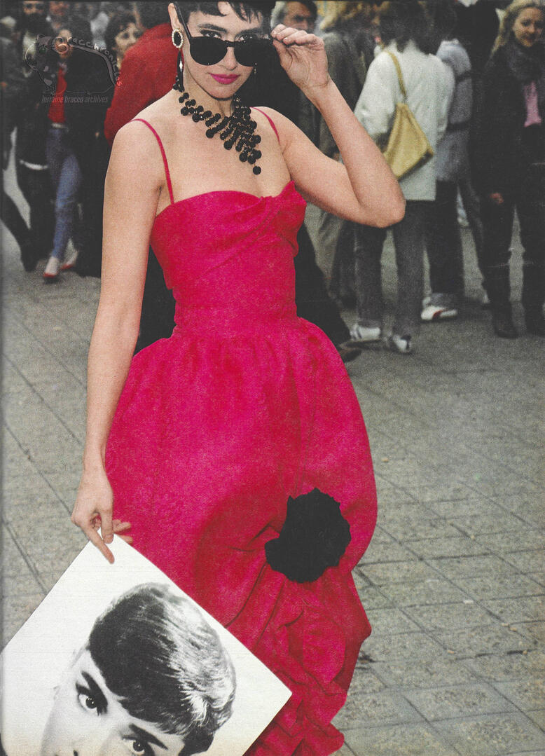 Continuation of Elle magazine spread showing Lorraine Bracco in a vibrant fuchsia strapless dress with a black flower detail. She wears sunglasses and a beaded necklace while holding a vintage portrait of Audrey Hepburn.