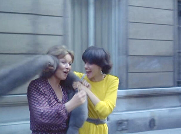 11 Lorraine Bracco in a vibrant yellow dress laughing on a street with a co-star in a scene from the 1979 French comedy movie Duos sur canapé.