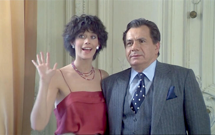 5 Lorraine Bracco waving in a red top alongside a man in a grey pinstripe suit during a scene from the 1979 French comedy movie Duos sur canapé.