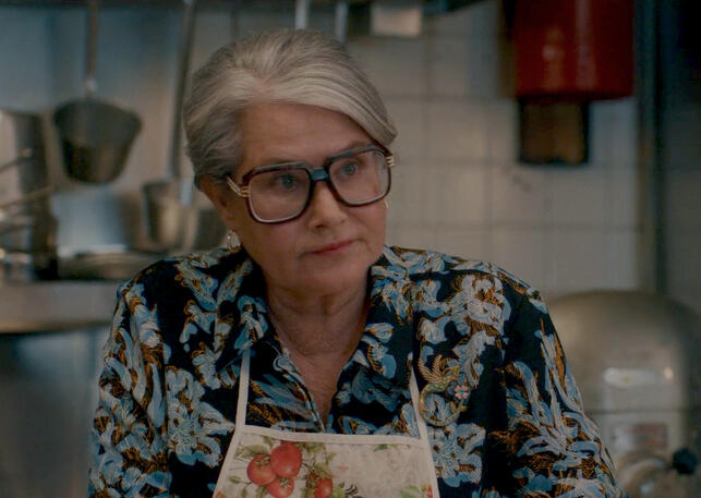 2 Lorraine Bracco wearing an apron and giant glasses in a kitchen scene from the film Nonnas.