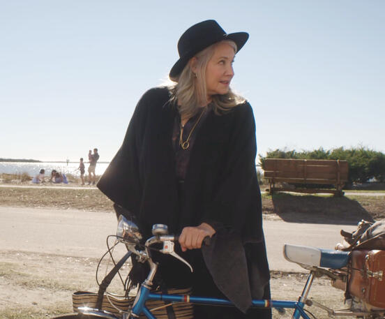 7 Lorraine Bracco pushing a bike outdoors in a black hat and cloak in Monster Summer.