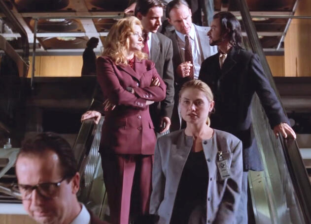 11 Lorraine Bracco in a maroon suit standing on an escalator with a group in a Hackers movie scene.