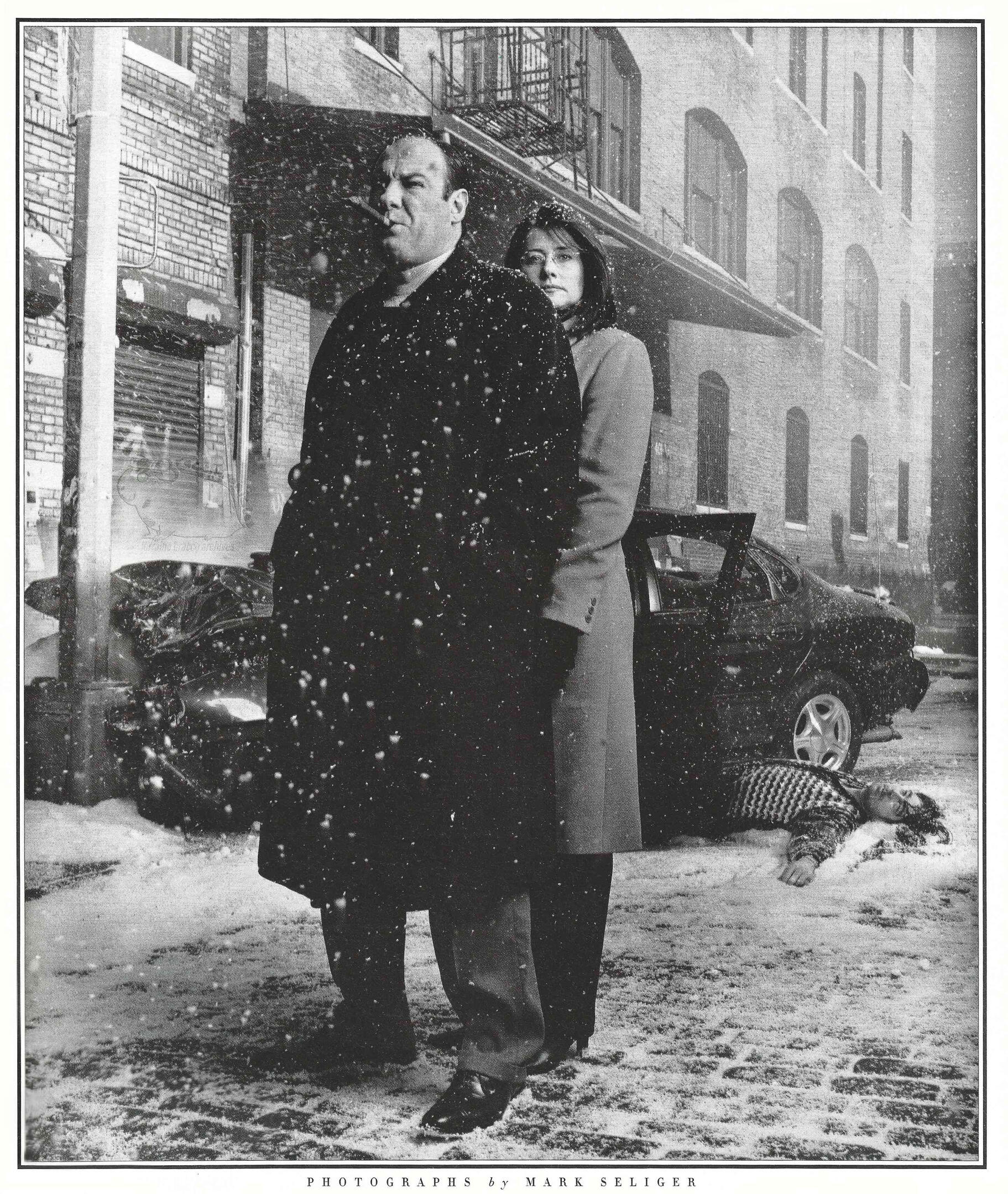 Cinematic black and white Rolling Stone magazine photograph by Mark Seliger featuring James Gandolfini smoking a cigar in a winter coat next to Lorraine Bracco. They stand on a snowy city street with a crashed car and a body lying in the snow behind them.