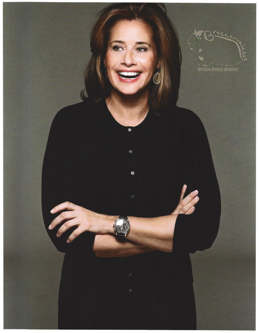 Portrait of actress Lorraine Bracco from the 'Bobbi Brown, Living Beauty' feature. She is smiling with her arms crossed, wearing a black button-up cardigan and a black watch against a grey background.